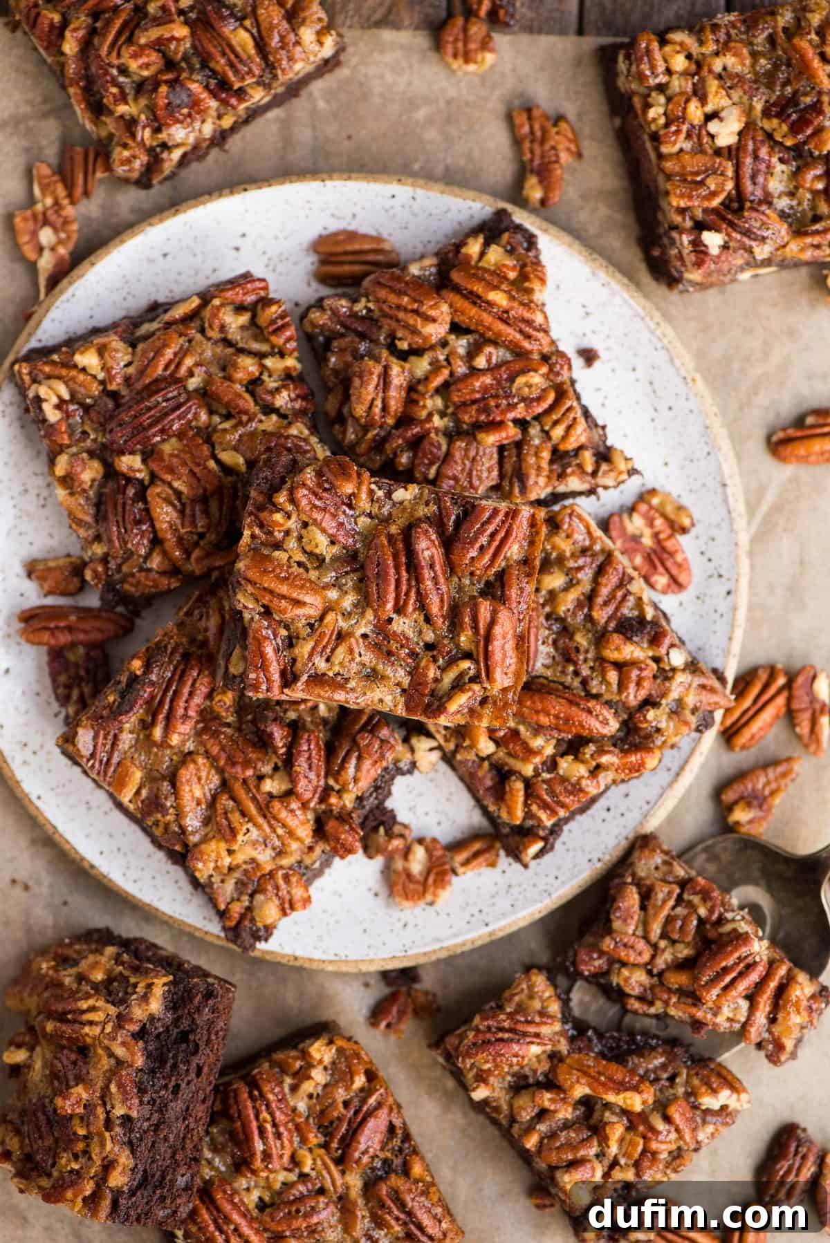 Stacked Pecan Pie Brownies on a white plate against a rustic wooden backdrop. The top brownie has a perfect crackly crust and visible chunks of pecan, emphasizing the two distinct layers.
