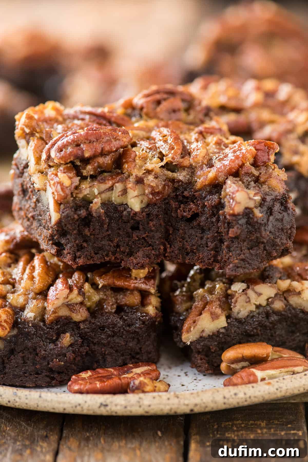 Two Pecan Pie Brownies stacked on a white plate, with a bite taken out of the top brownie, revealing its fudgy chocolate interior and the gooey pecan pie layer.
