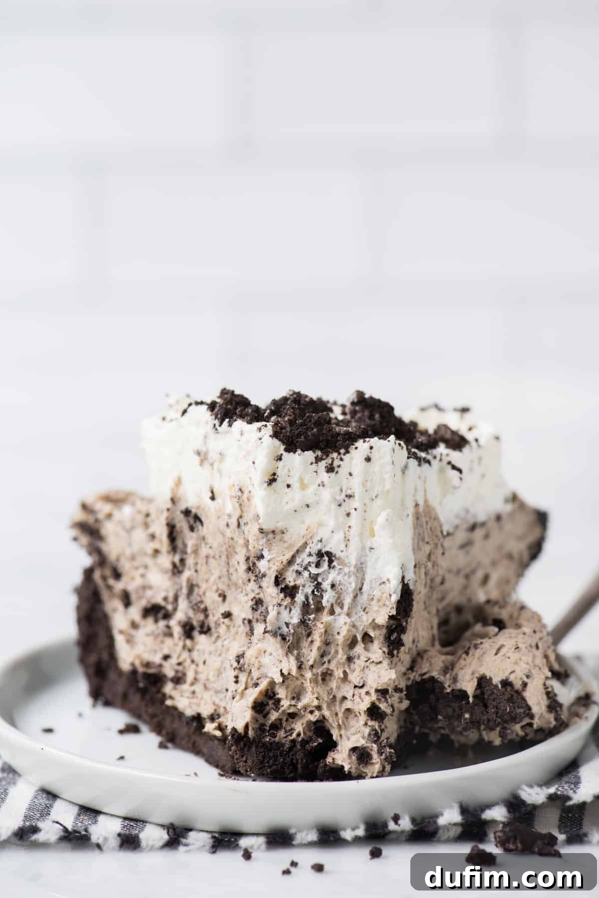 A perfectly sliced Oreo pudding pie on a white plate, showcasing its layered texture with a rich dark crust, creamy vanilla filling, and a dollop of whipped topping, set against a crisp white linen.