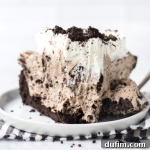 A tempting slice of chocolate and cream dessert featuring a crumbly Oreo cookie crust, a light chocolate mousse filling, and a generous swirl of whipped cream topping—a classic Oreo pudding pie—served on a pristine white plate beside a checkered napkin.