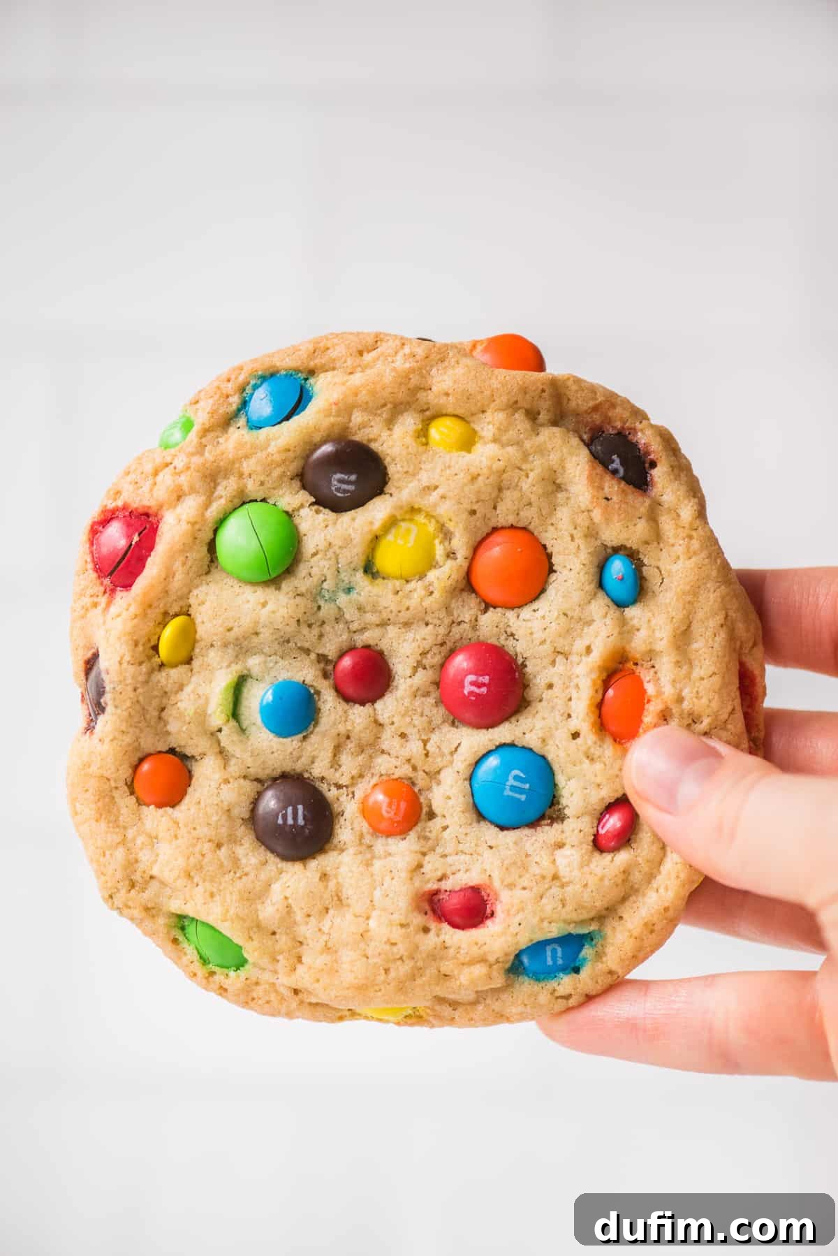 Chewy M&M Cookie Recipe 4 hand holding M&M cookie on white background, showcasing its soft texture