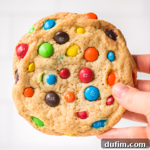 Chewy M&M Cookie Recipe 8 A hand is holding a large, round M&M cookie filled with colorful candy-coated chocolate pieces against a light background.