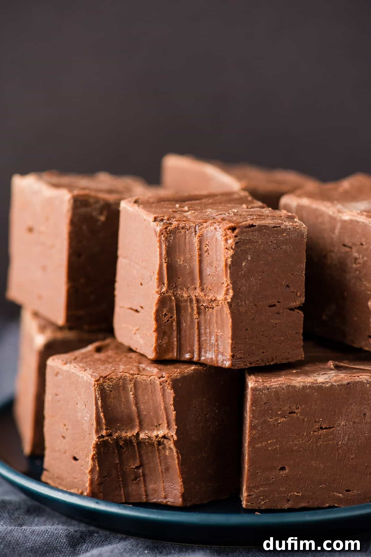 Stacked squares of tall, thick homemade chocolate fudge on a navy blue background, highlighting its rich, dark color and smooth texture.