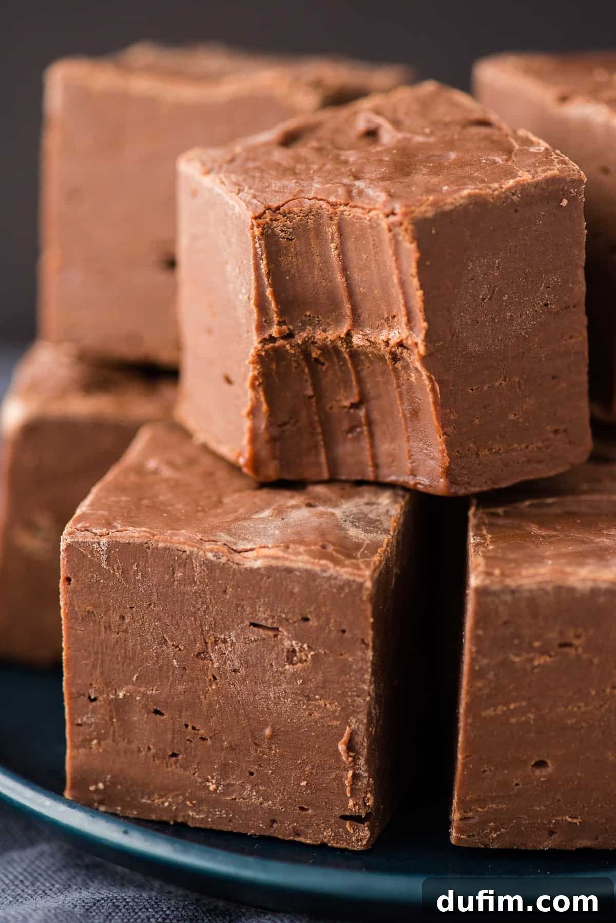 Multiple pieces of homemade chocolate fudge stacked artfully on a navy blue surface with a dark background, emphasizing their height and rich appearance.