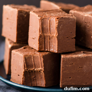 A close-up of several thick, creamy chocolate fudge squares stacked on a dark plate, with visible bite marks on the top pieces, perfectly highlighting their smooth and rich chocolate fudge texture.