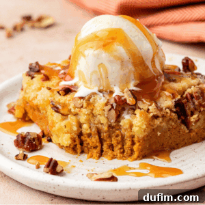 A square slice of pumpkin dump cake, beautifully plated and topped with a generous scoop of vanilla ice cream, chopped pecans, and a luscious drizzle of caramel sauce. A bite has been taken, revealing the moist interior.