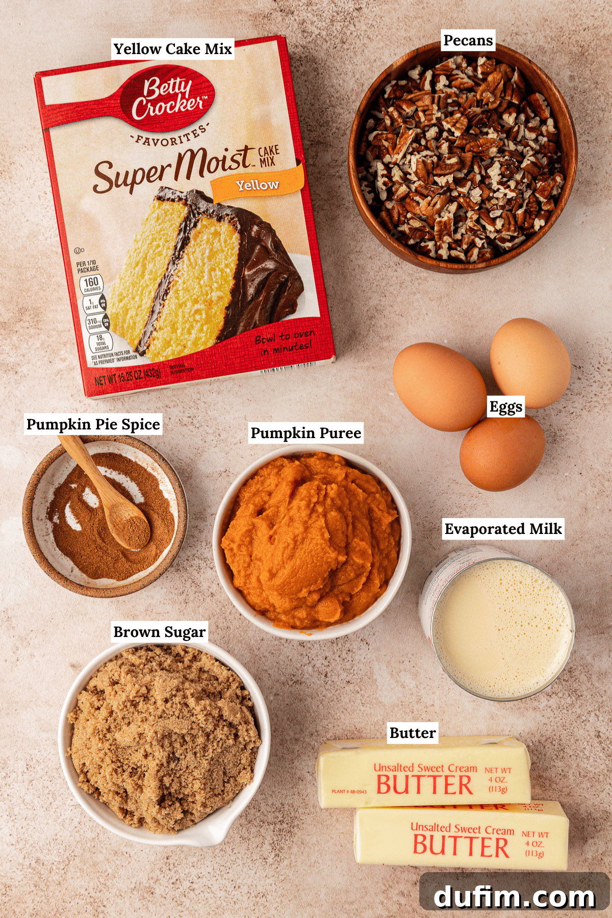 Effortless Autumn Pumpkin Dump Cake 6 All the essential ingredients for a pumpkin dump cake recipe beautifully arranged on a white surface: a box of yellow cake mix, a bowl of chopped pecans, a carton of eggs, a can of evaporated milk, sticks of unsalted butter, a bowl of brown sugar, a can of pumpkin puree, and a jar of pumpkin pie spice—each clearly labeled and ready for use.