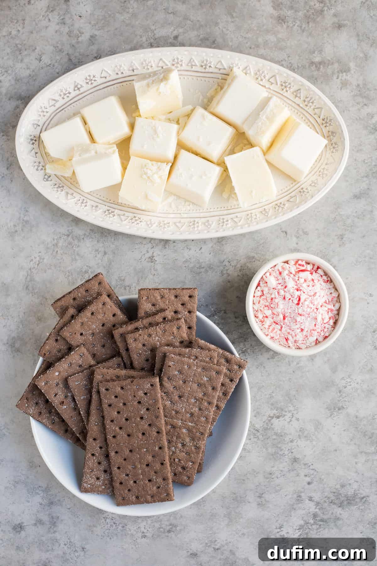 A flat lay photograph showcasing the three main ingredients: a box of chocolate graham crackers, a package of vanilla almond bark, and a pile of whole candy canes, ready for making chocolate peppermint bark.