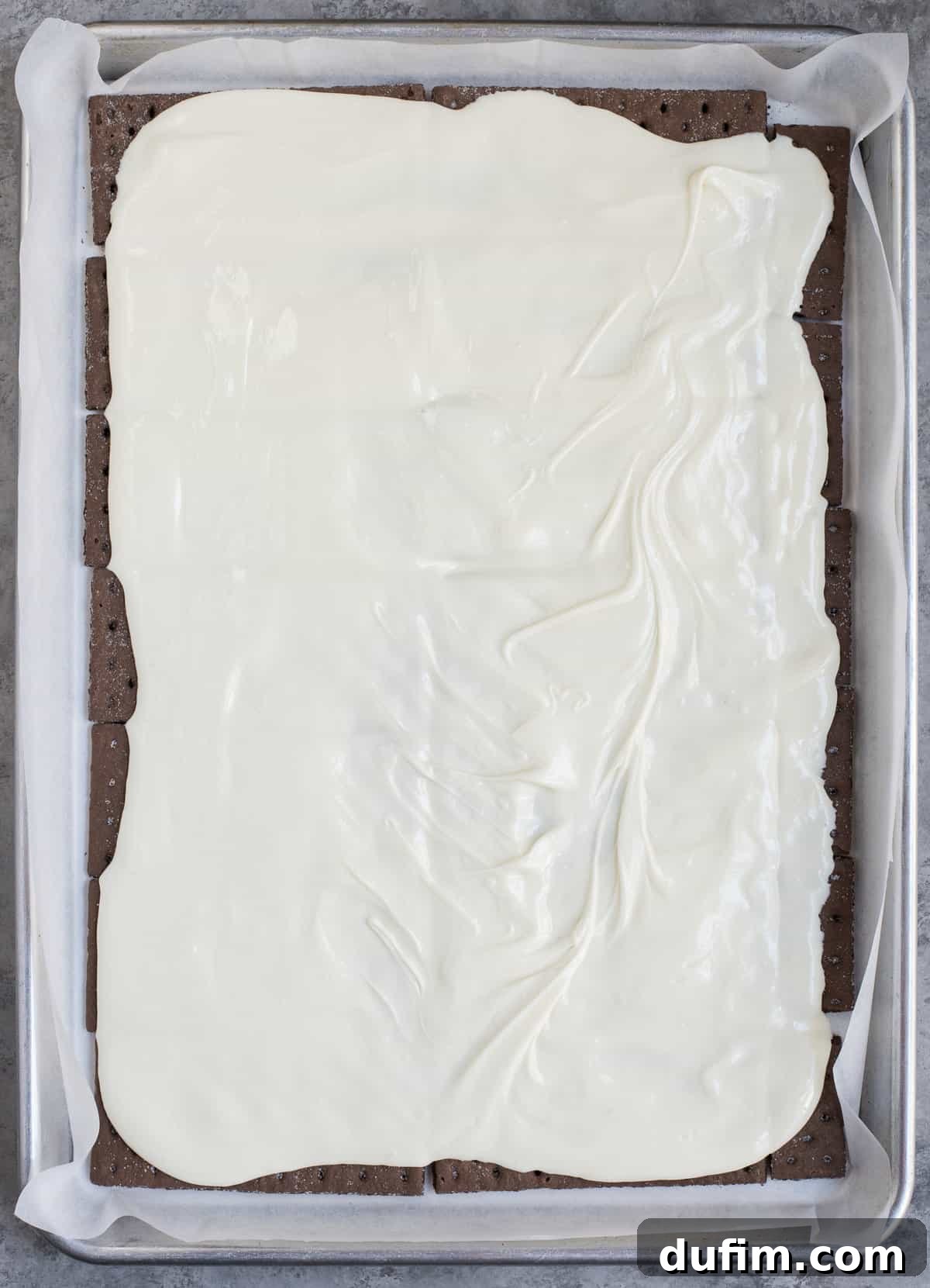 Smooth, melted vanilla almond bark spread evenly over a single layer of chocolate graham crackers on a baking sheet, ready for the peppermint topping.
