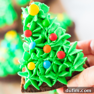 A hand holds a festive Christmas tree brownie, decorated with bright green frosting and colorful round candies resembling ornaments.
