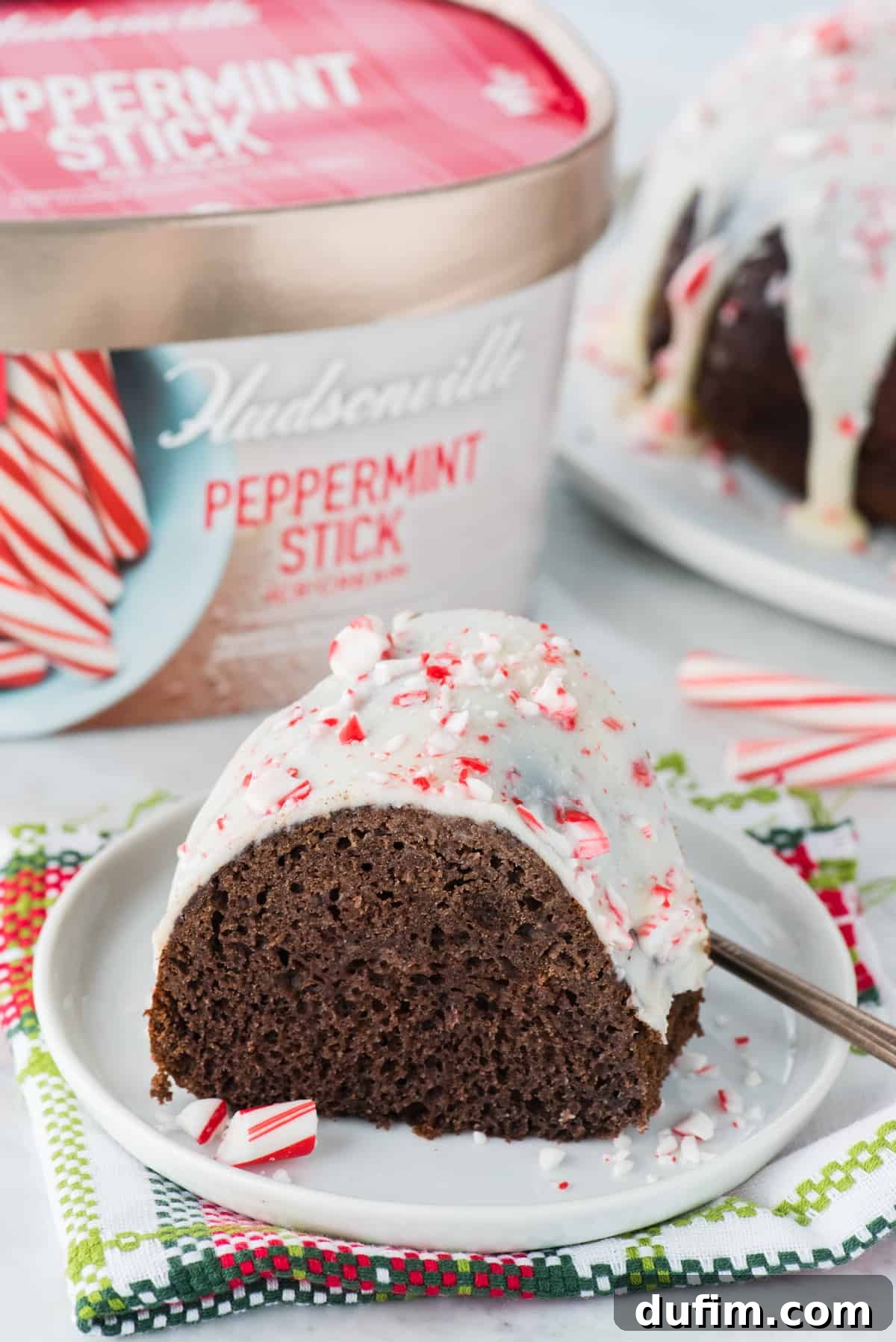 A delectable slice of chocolate peppermint bundt cake on a white plate, with a container of peppermint stick ice cream blurred in the background, hinting at the key ingredient.