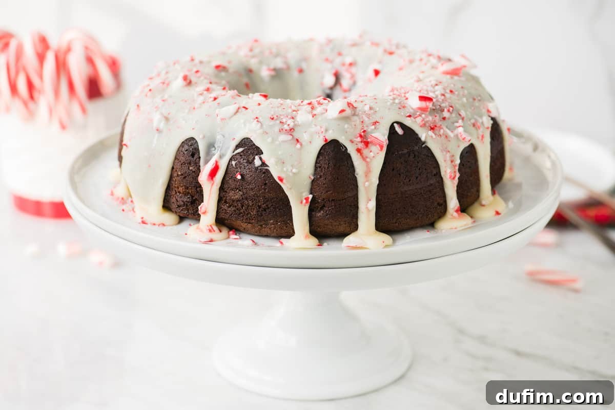 A stunning whole chocolate bundt cake, elegantly drizzled with white chocolate glaze and adorned with crushed candy cane pieces, presented on a pristine white cake stand.