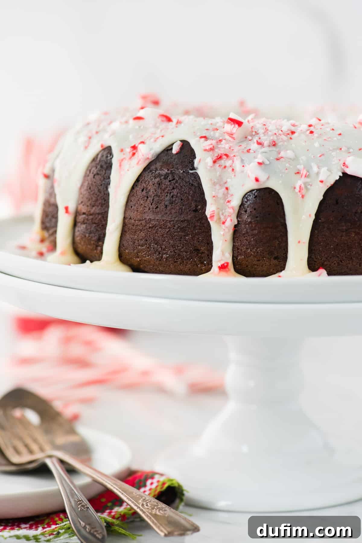 A full view of the chocolate peppermint bundt cake, beautifully glazed with white chocolate and sprinkled with crushed candy canes, proudly displayed on a white cake stand.
