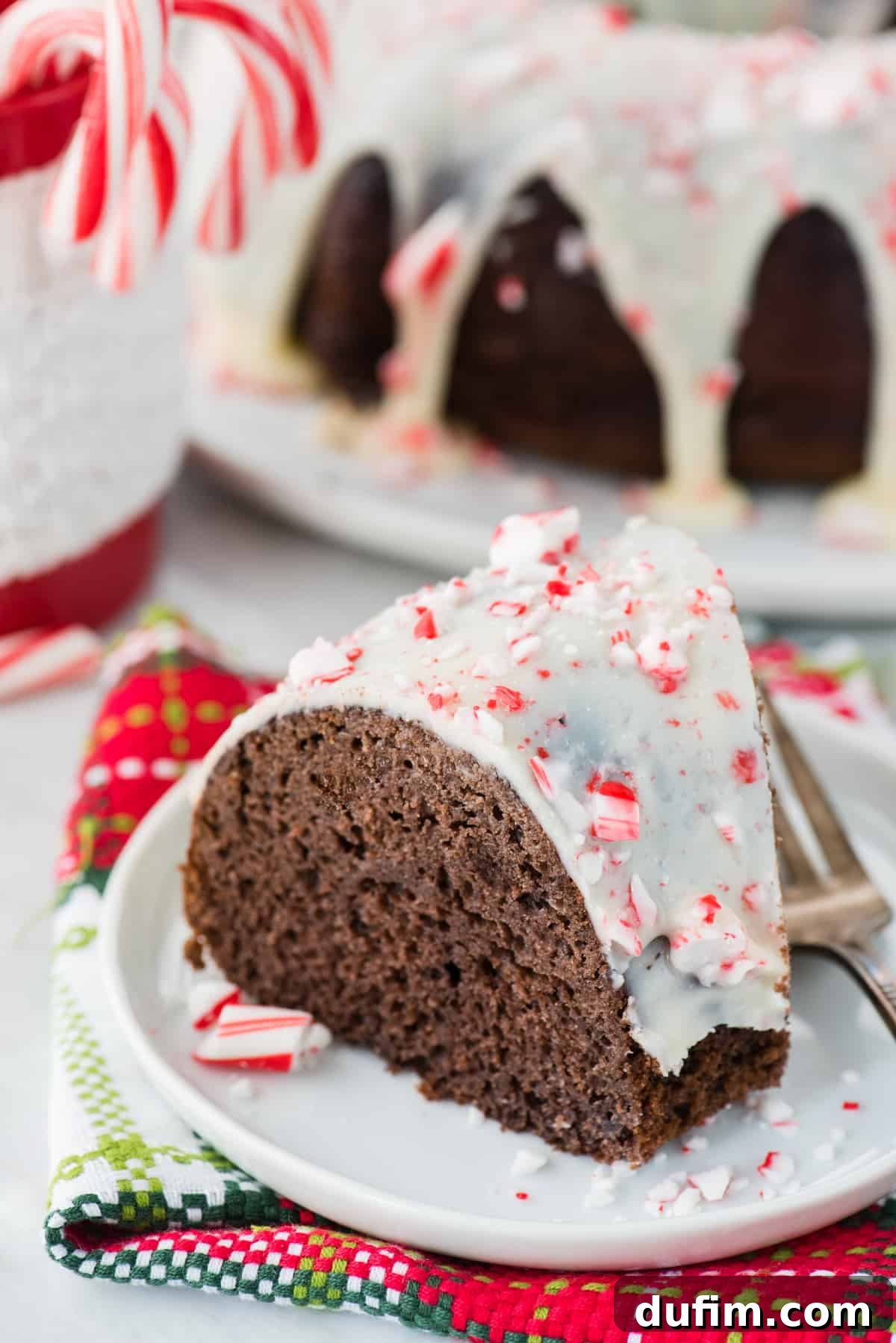 A close-up of a slice of chocolate peppermint bundt cake, featuring its moist interior, white glaze, and candy cane pieces, with more candy canes in the background.