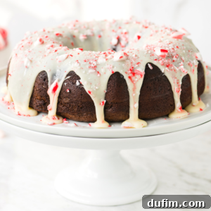 A beautifully decorated chocolate peppermint bundt cake with white icing and crushed peppermint candy on top, placed on a white cake stand against a softly lit background, ready to be enjoyed.