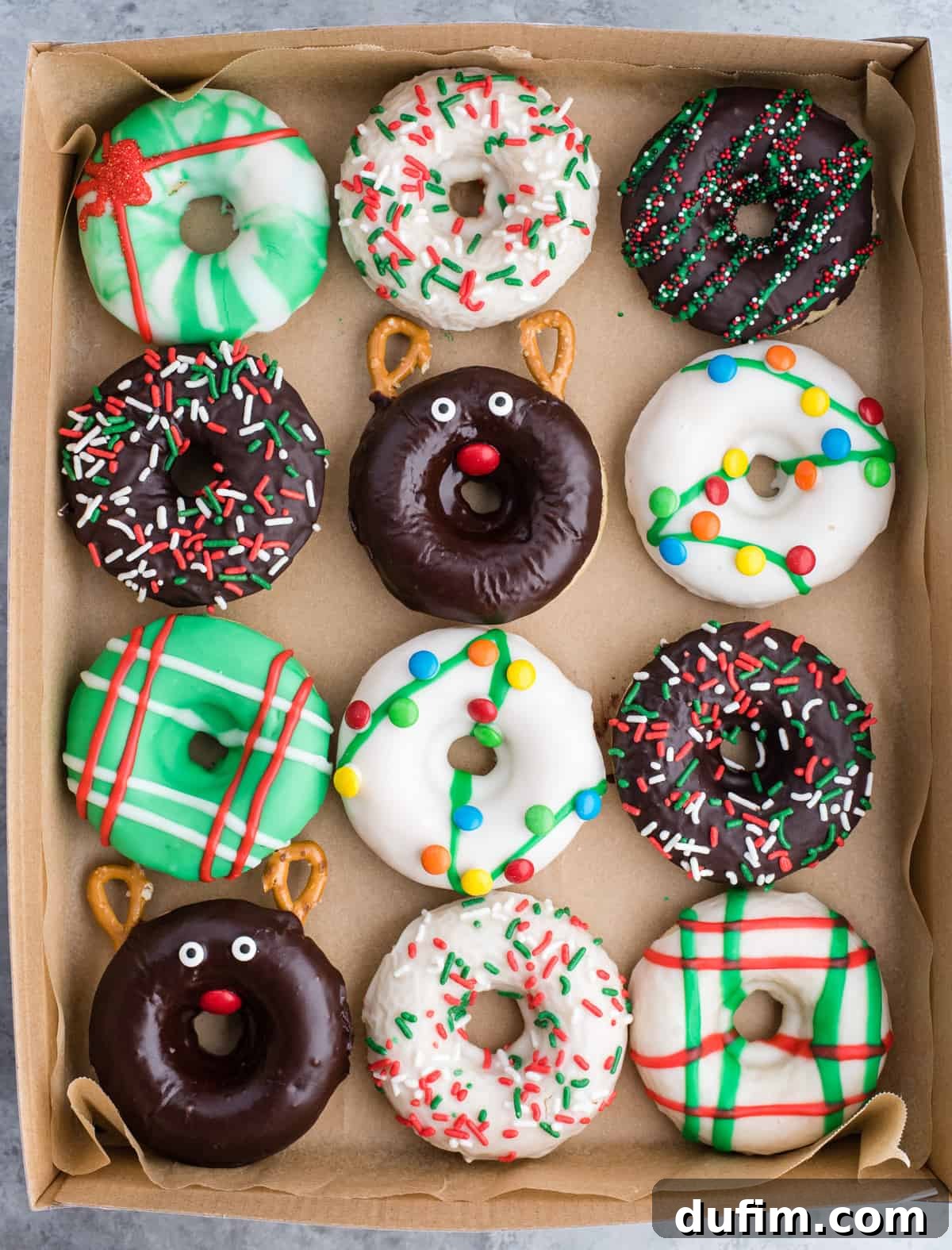 A box of twelve beautifully decorated Christmas donuts, arranged neatly, showcasing various festive designs like sprinkles, plaid, and reindeer, ready for gifting or sharing.