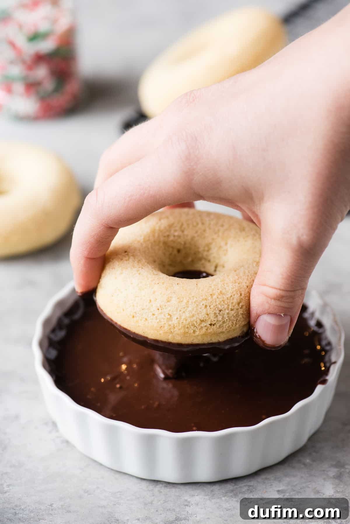 A person dipping a freshly baked donut into a bowl of glossy white glaze, with excess glaze gracefully dripping back into the bowl.
