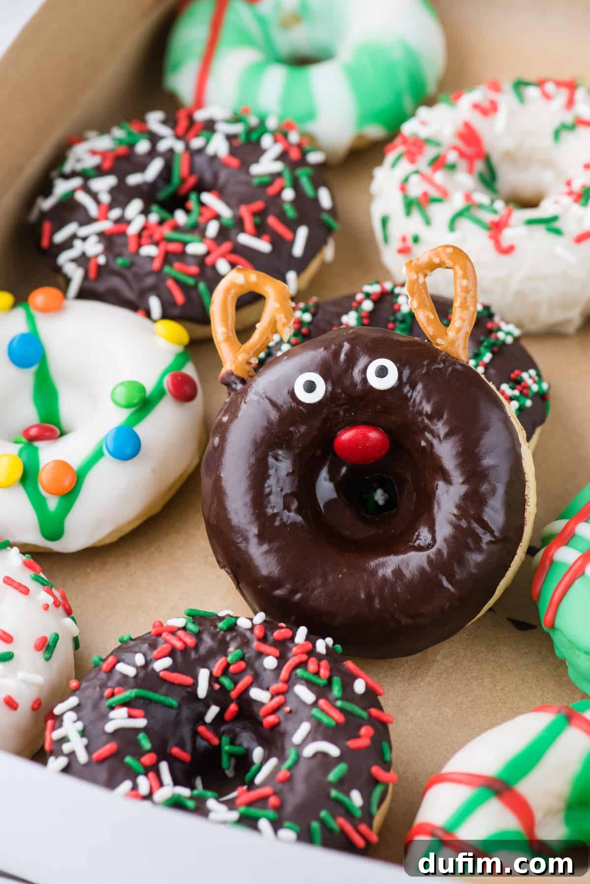 A close-up view of a charming chocolate reindeer donut, meticulously decorated with pretzels for antlers and candy eyeballs, with other festive donuts visible in a donut box, ready for holiday gifting or sharing.