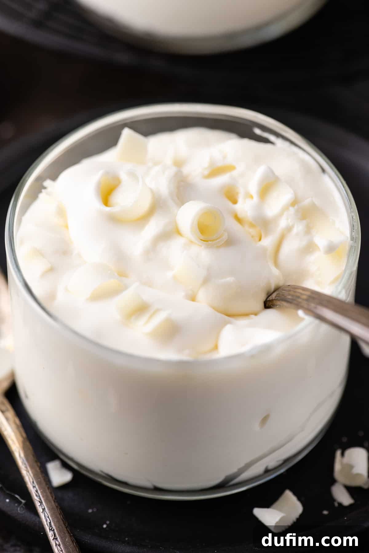 Indulgent White Chocolate Mousse 4 A shining metal spoon nestled in a glass cup brimming with smooth white chocolate mousse, set against a dark, inviting background.