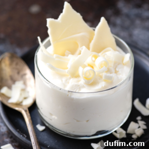 A glass cup filled with creamy white chocolate mousse, topped with delicate curls and shards, sits on a dark plate next to a gold spoon. White chocolate pieces are scattered around.