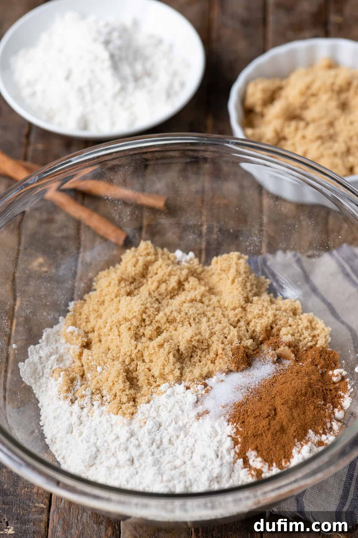 A glass bowl holds the dry ingredients for crumb topping: flour, brown sugar, and cinnamon, arranged on a warm wooden background.