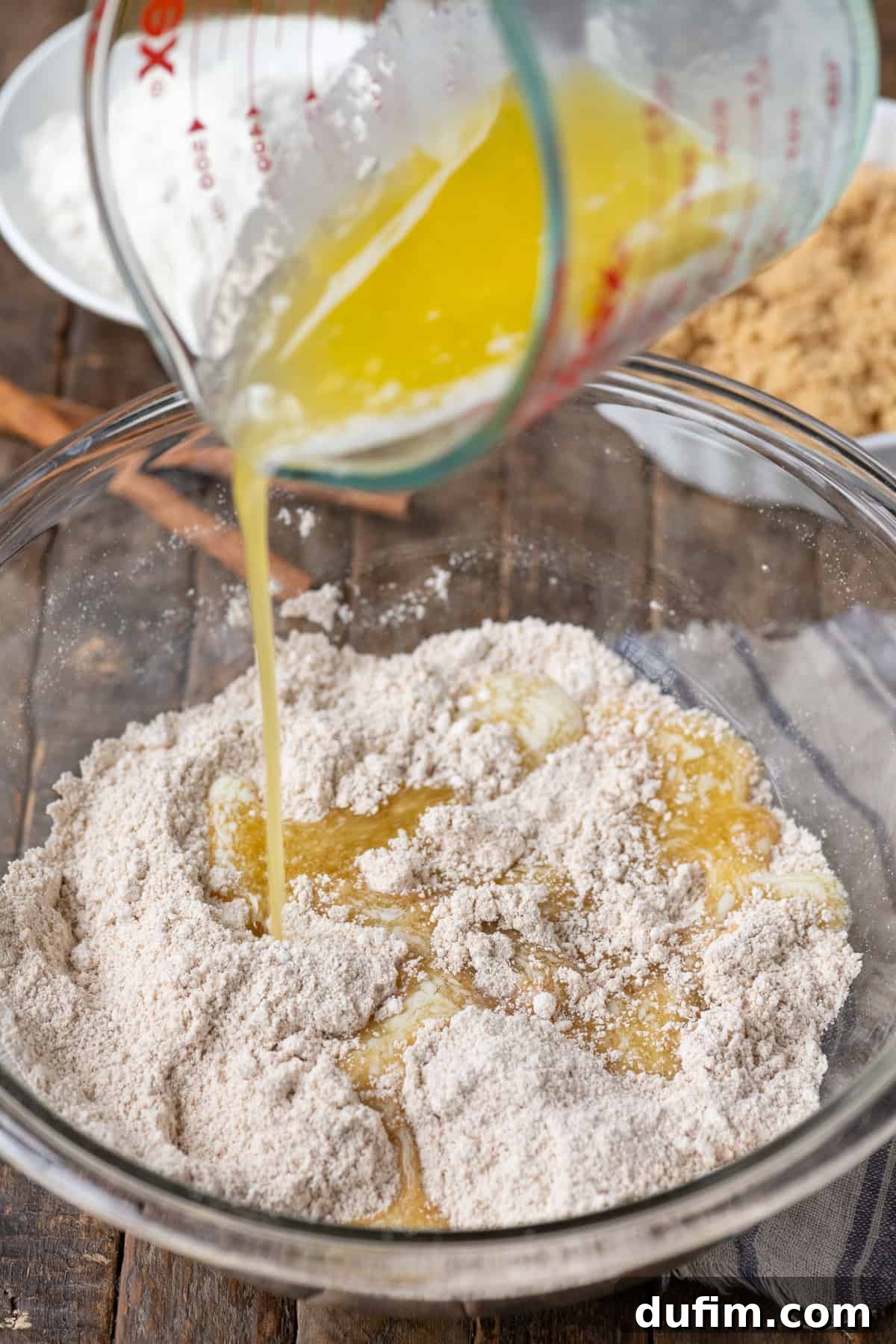 Melted butter being gently poured into a glass bowl containing the dry crumb topping mixture, set on a wooden surface.