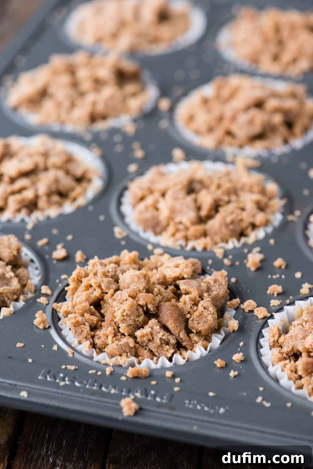 A metal muffin pan is generously filled with muffin batter, adorned with a high pile of crumb topping, awaiting baking.