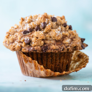 A close-up of a chocolate chip muffin with a golden streusel topping, partially unwrapped from its brown paper liner, set against a light blue background.