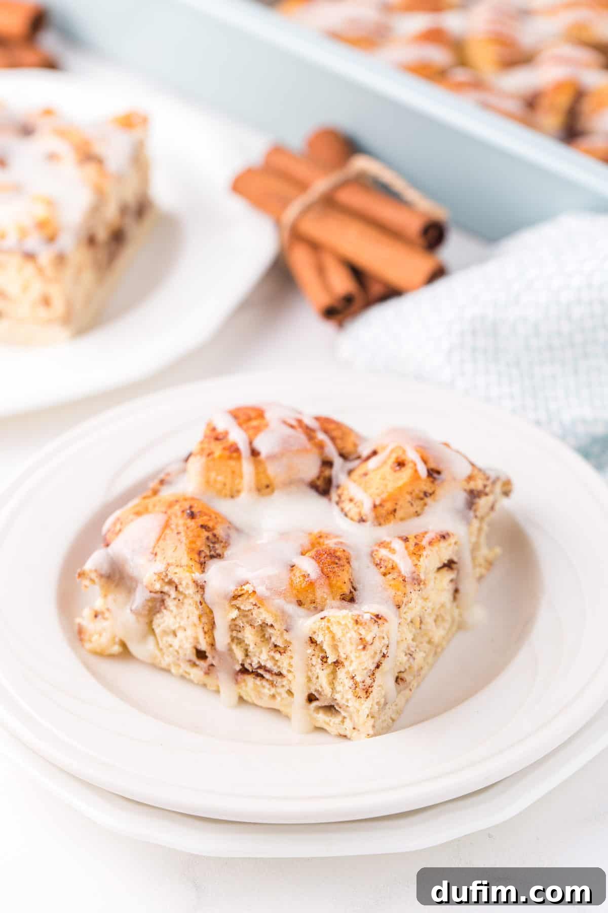 A perfectly baked slice of cinnamon roll casserole, generously topped with a sweet white icing, presented on a pristine white plate.