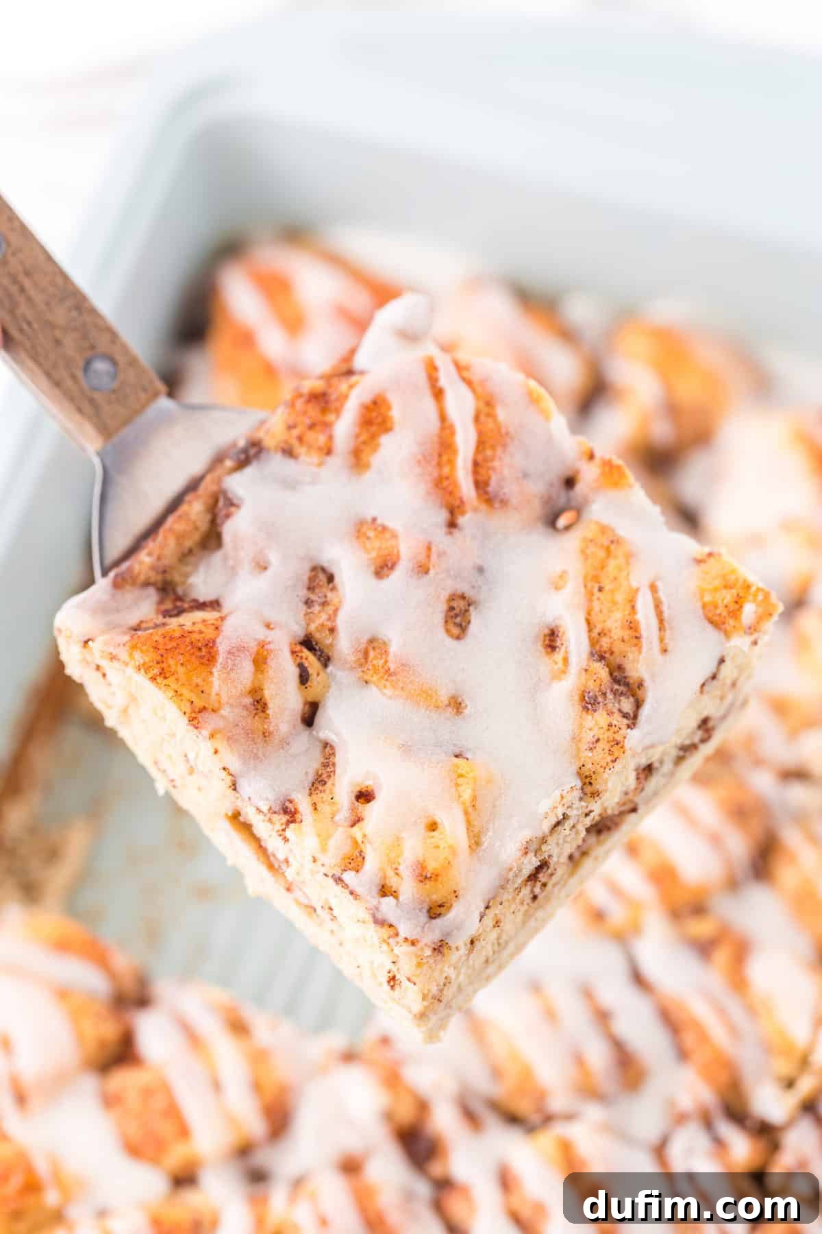 A serving of cinnamon roll casserole being lifted from the pan with a metal spatula, showing its gooey texture and golden crust.