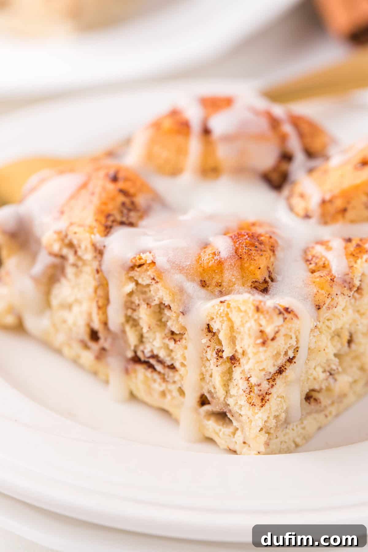 A delightful slice of cinnamon roll casserole, generously coated with white icing, sitting on a clean white plate, ready to be eaten.