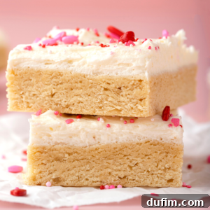 Two thick valentines day sugar cookie bars stacked, topped with creamy white frosting and decorated with pink, red, and white sprinkles, rest on crinkled parchment paper against a pink background—perfect for Valentines cookie ideas.