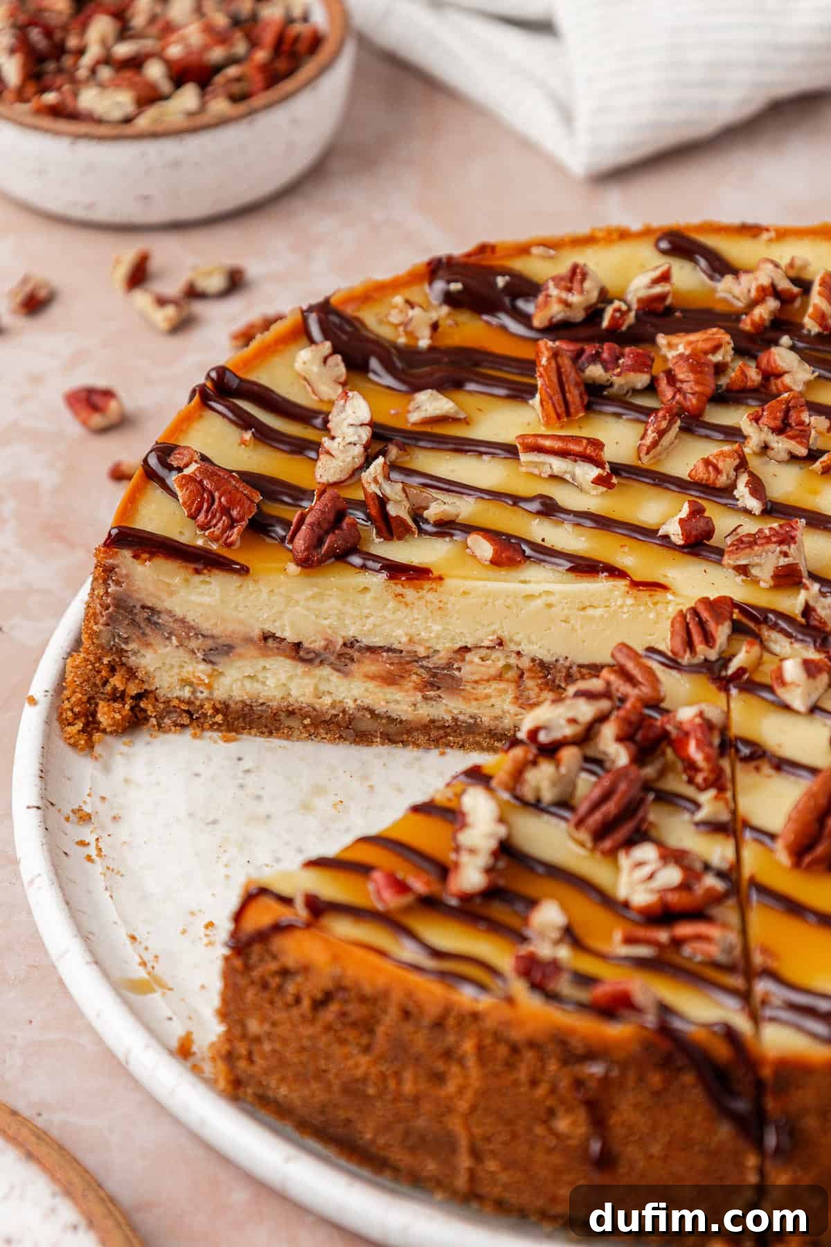 A caramel and chocolate-drizzled cheesecake topped with chopped pecans sits on a white plate, with one slice removed. Crumbs and extra pecans are scattered around the plate and table.