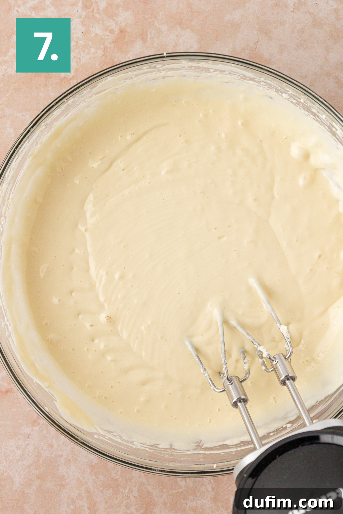 A glass bowl filled with smooth cake batter being mixed by an electric hand mixer, viewed from above. A green square with the number 7 is in the top left corner.