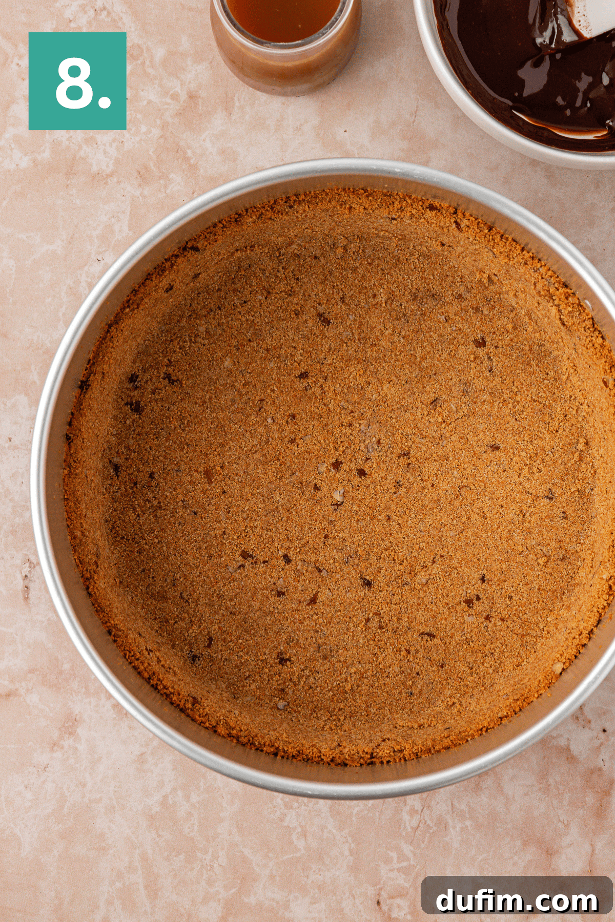 A round metal pie pan filled with a pressed graham cracker crust sits on a marble surface. The corner shows a bowl of melted chocolate, a small cup, and a green square with the number 8 in white.