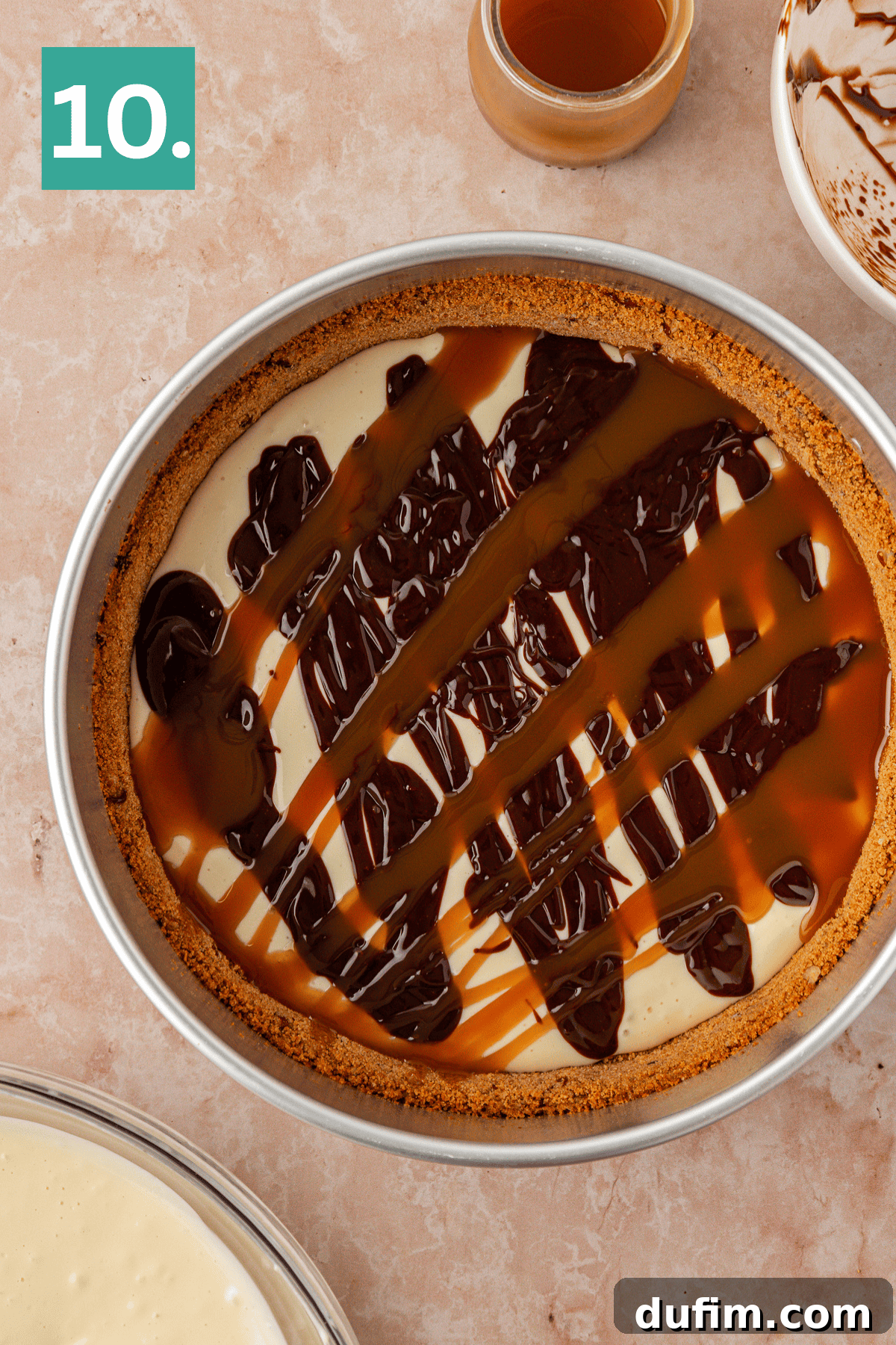 A round cheesecake in a pan is topped with drizzles of chocolate and caramel sauce. A jar of caramel and a bowl with chocolate remnants are nearby. A green box with the number 10 is in the top left corner.