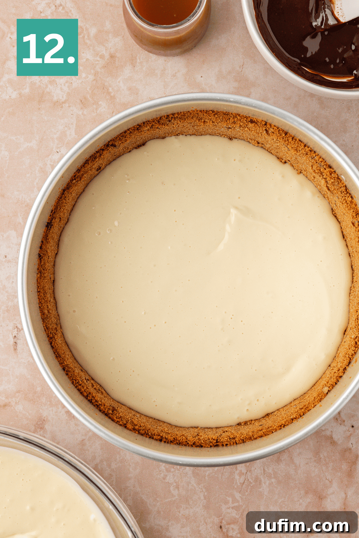 A cheesecake in a round pan with a graham cracker crust, unbaked and smooth on top, sits on a light countertop. A small bowl of caramel and a bowl of chocolate are nearby. A green box with the number 12 is in the corner.