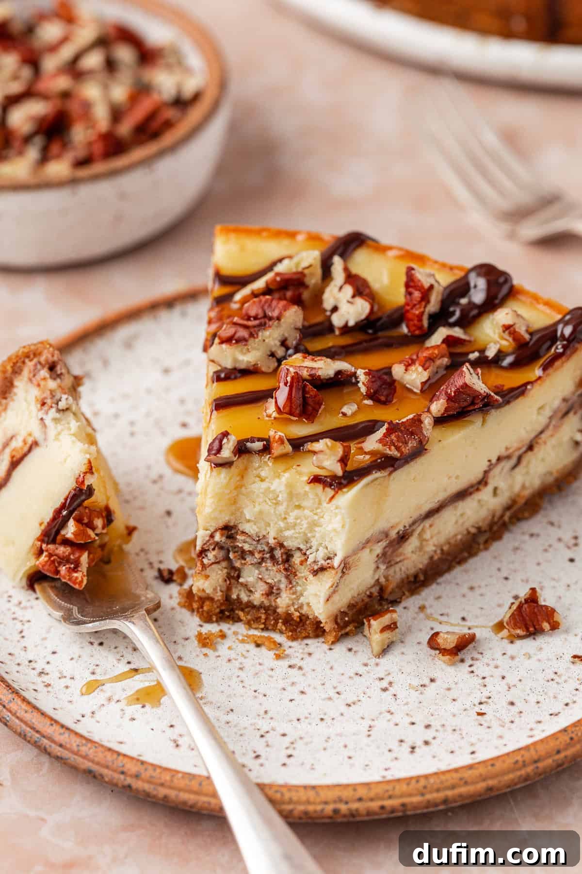 A slice of creamy cheesecake topped with chopped pecans, caramel, and chocolate drizzle sits on a speckled plate. A fork holds a bite of the cheesecake, and more dessert is in the background.