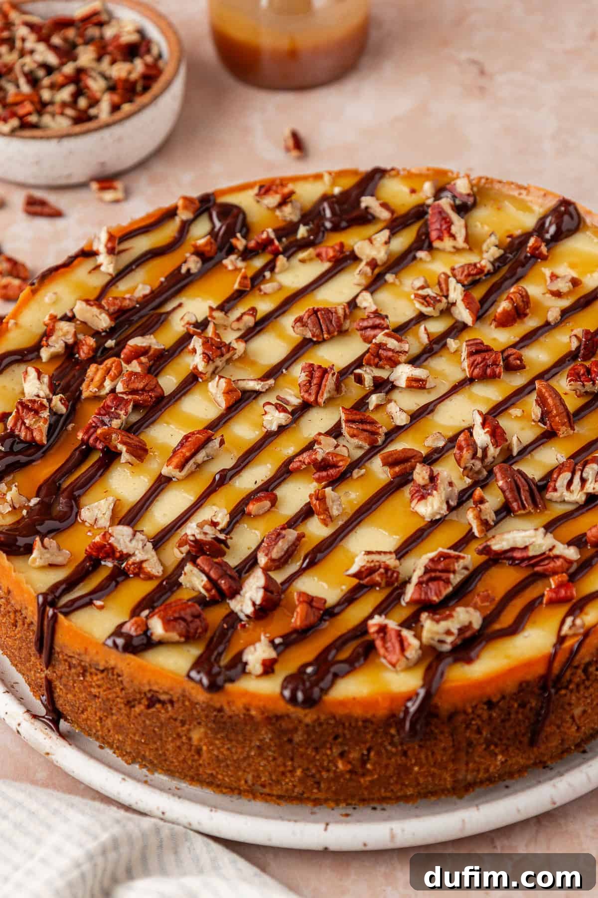 A cheesecake topped with chopped pecans, caramel sauce, and chocolate drizzle, displayed on a round white plate. A bowl of extra pecans and a jar of caramel sauce are in the background.