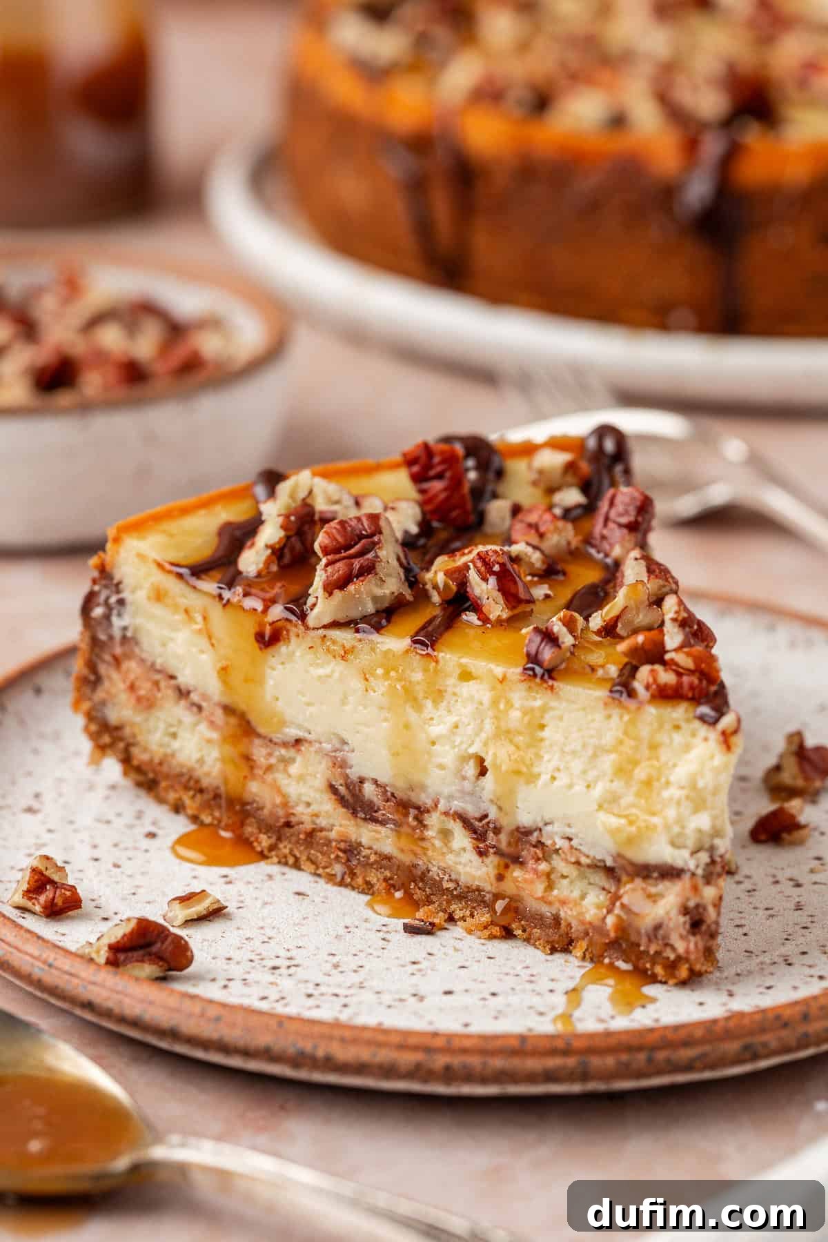A slice of cheesecake topped with chopped pecans and caramel drizzle sits on a speckled plate, with more pecans and a whole cheesecake visible in the background.