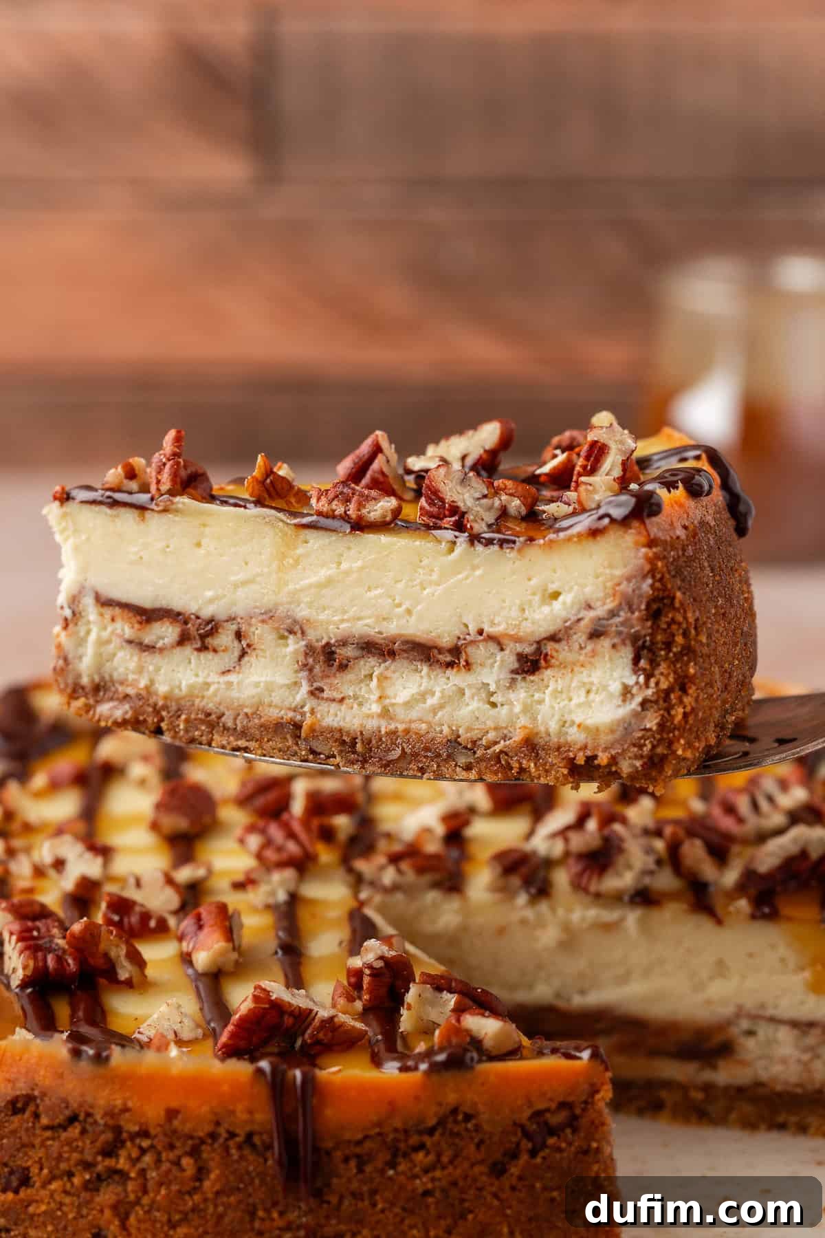 A close-up of a slice of cheesecake with a graham cracker crust, chocolate layer, and chopped pecans on top. The slice is being lifted from the whole cake in the background.