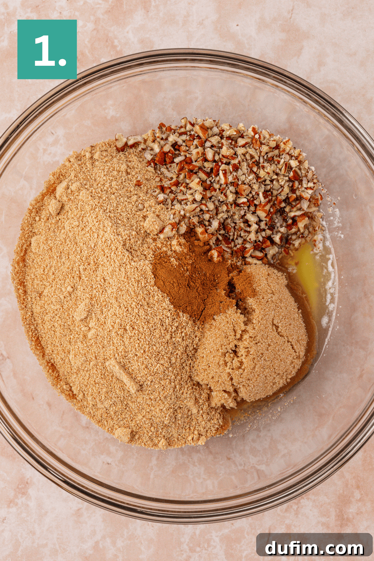A glass bowl filled with graham cracker crumbs, chopped pecans, brown sugar, cinnamon, and melted butter, viewed from above. A green box with the number 1 is in the top left corner.