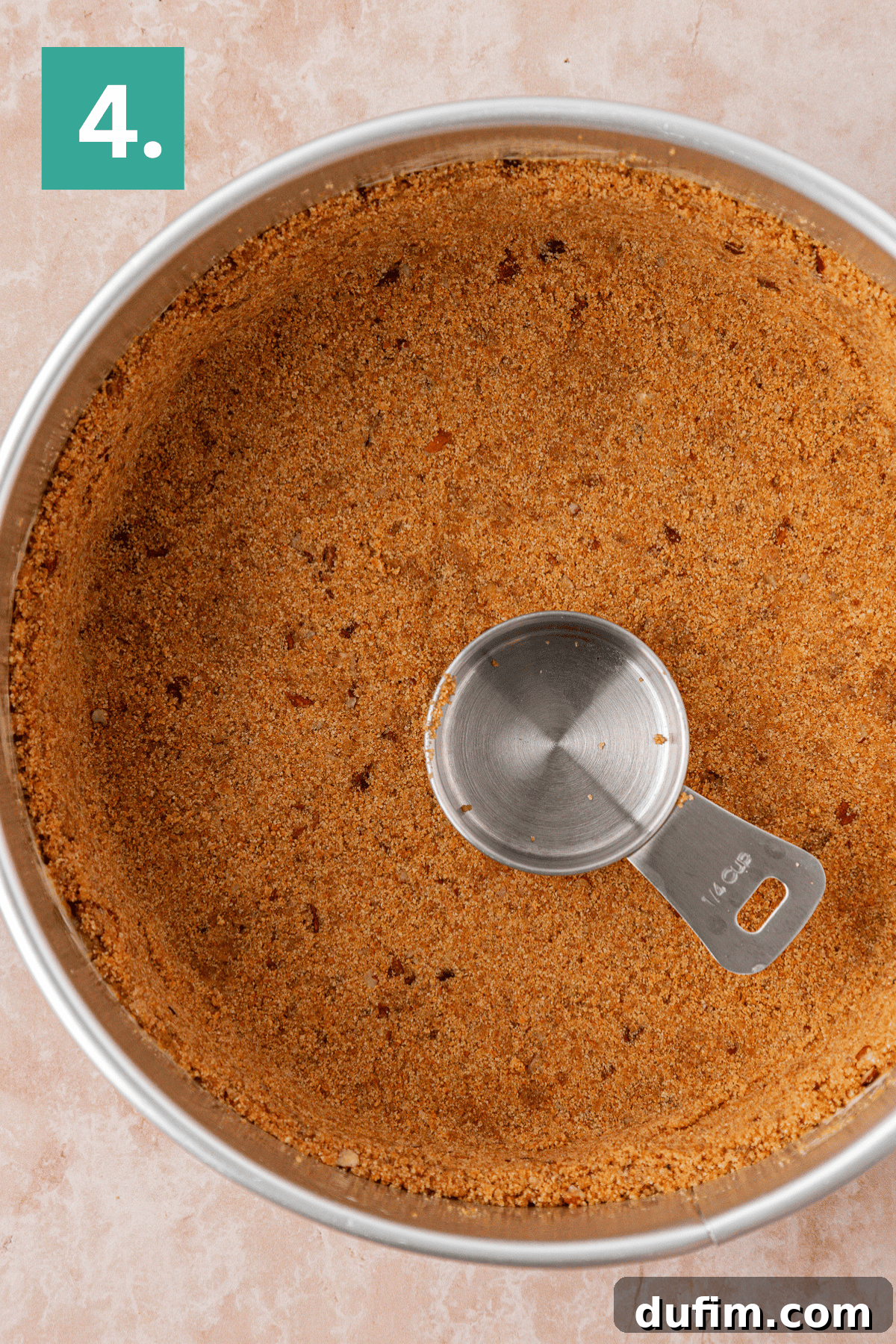 A round metal pan filled with an even layer of crumb crust and a metal measuring cup pressing the crust. A green box with the number 4 is in the top left corner.