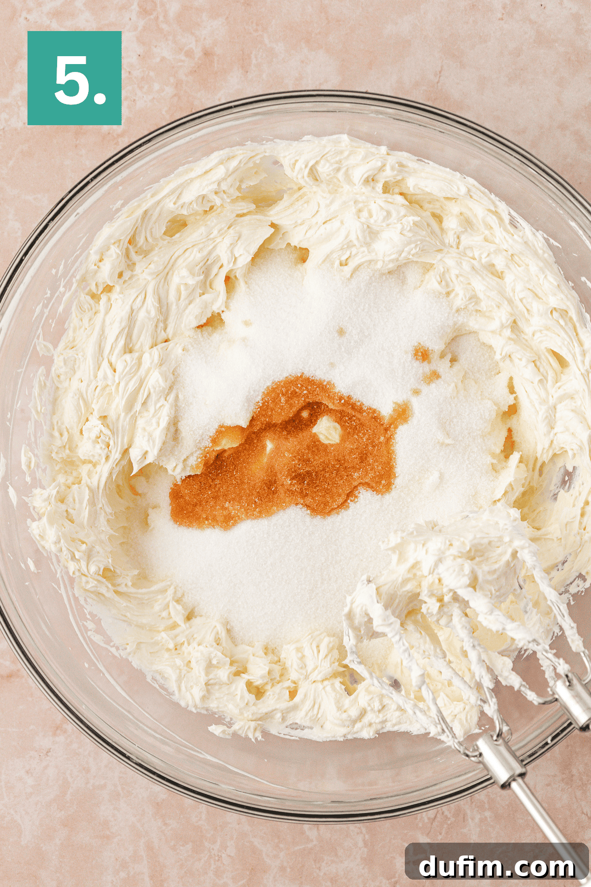 A glass mixing bowl with whipped cream cheese, sugar, and vanilla extract, shown with electric mixer beaters. A green square with the number 5 appears in the top left corner.