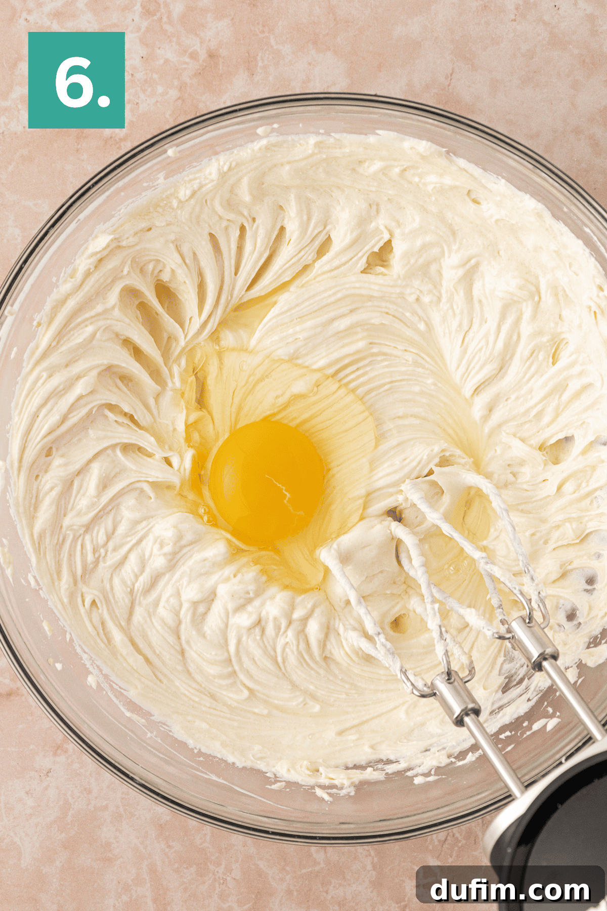 A glass bowl with creamy batter and a whole egg in the center, next to an electric hand mixer with beaters. A green box with the number 6 is in the top left corner.