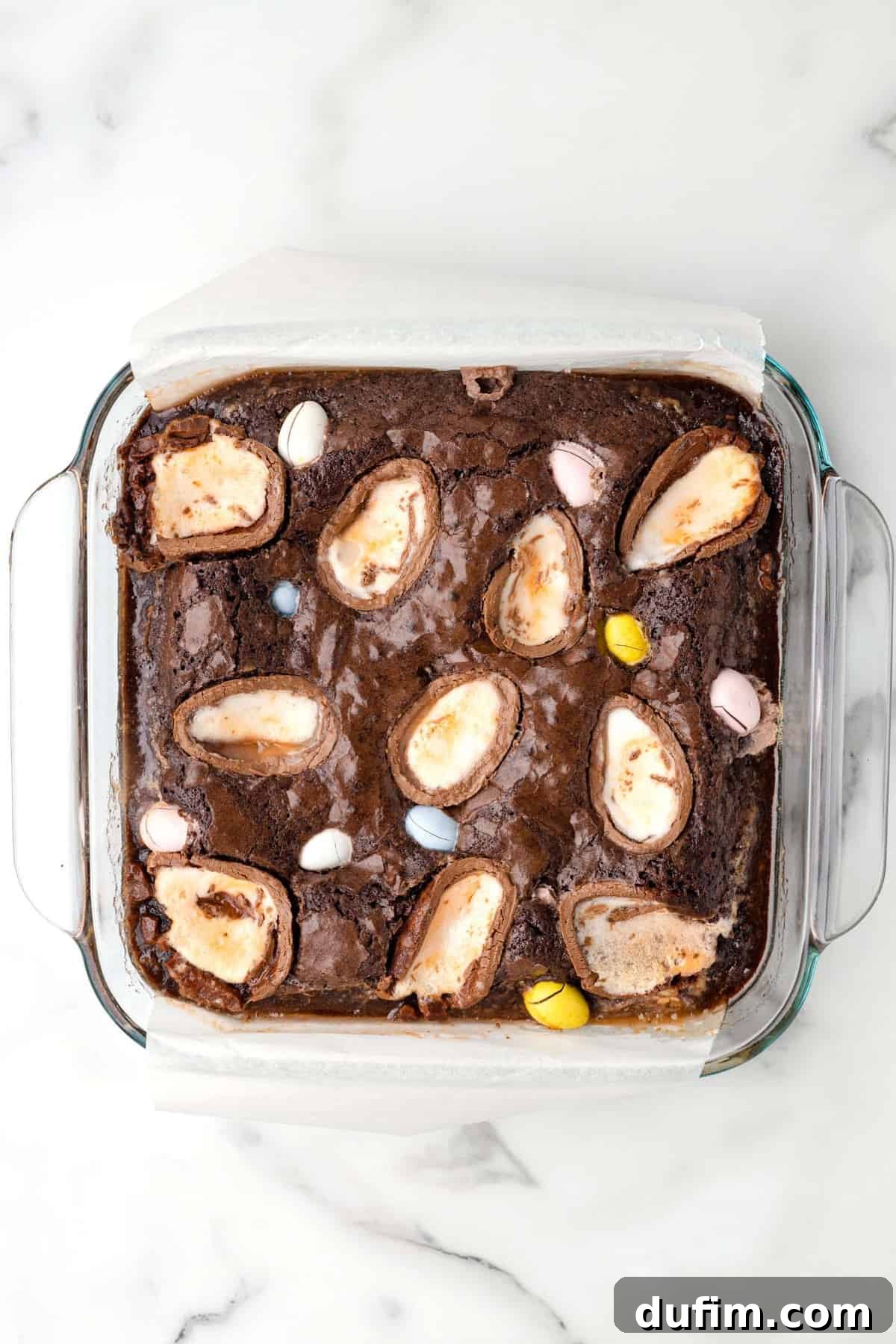 A top-down view of freshly baked Cadbury Creme Egg Brownies in a clear glass baking dish, adorned with colorful mini eggs and melted Creme Eggs, set against a pristine white background.