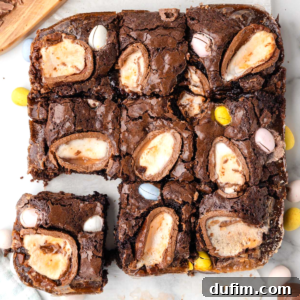 A batch of Cadbury Creme Egg Brownies cut into squares, each topped with a halved creme egg and small colorful chocolate eggs, displayed on a white surface. One brownie piece is separated from the batch, showcasing its rich, fudgy interior.