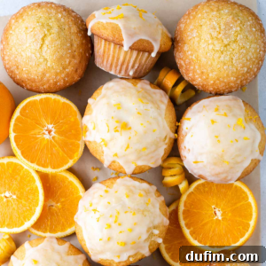 Delicate orange muffins topped with a sweet white glaze and fragrant orange zest, artfully arranged on parchment paper alongside whole and sliced fresh oranges, with two plain sugar-topped muffins in the background.