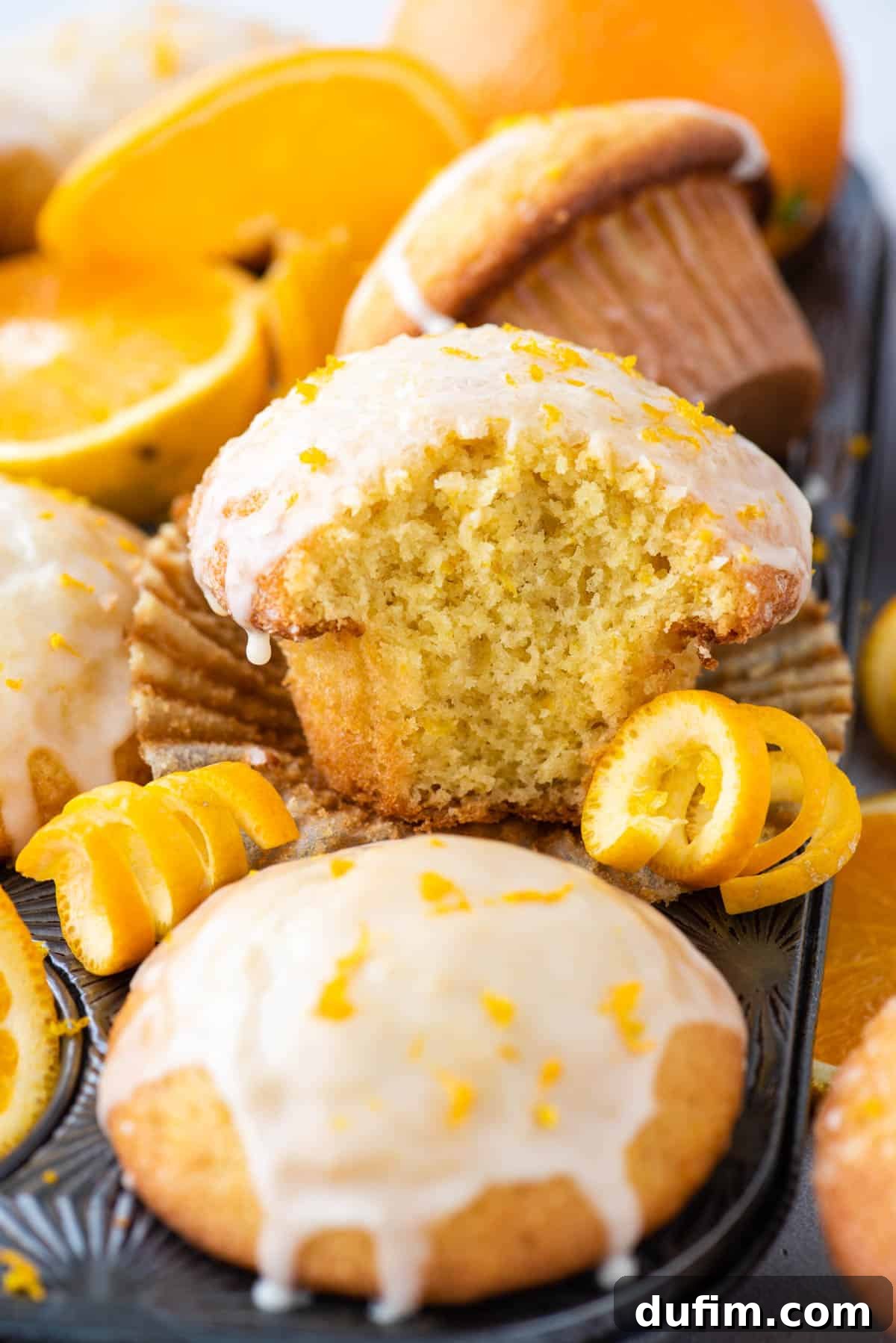 A perfectly baked orange muffin with a golden, domed top and a visible bite taken out, resting in a muffin pan, showing its moist and tender interior.