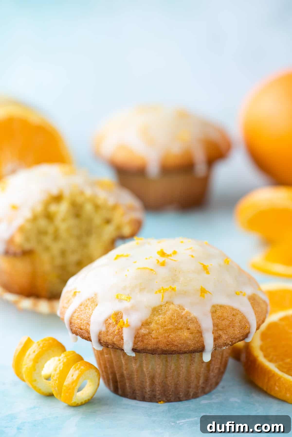 A single, perfectly baked orange muffin sits on a white plate, surrounded by more muffins and fresh orange slices on a rustic blue background, emphasizing its homemade appeal.